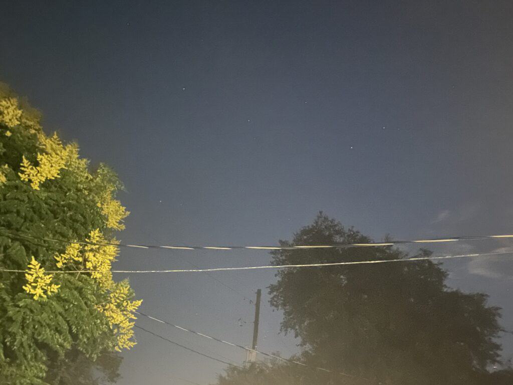 Small patch of open night sky framed by trees, a roofline, and power lines in a light-polluted backyard
